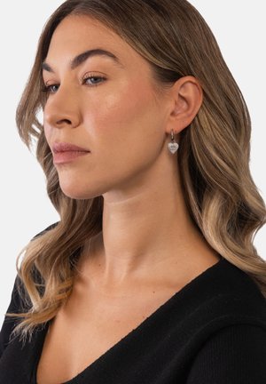 Frau mit hellbraunem, welligem Haar, die ein schwarzes Oberteil und silberne, herzförmige Ohrringe mit Sonnenstrahl-Design trägt, blickt leicht nach links.