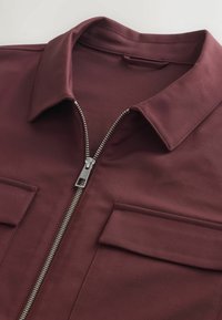 Burgundy jacket with a pointed collar, silver zipper, and two chest pockets. Made of smooth fabric with a sleek finish and minimal detailing.