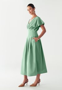 Vestido verde menta con un escote en V profundo, mangas cortas con vuelo y bolsillos laterales. Falda acampanada con pliegues. Combinado con tacones marrones con estampado.