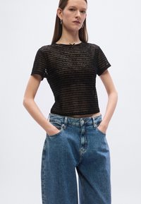 Juoda trumpomis rankovėmis palaidinė su permatomu audiniu ir pasikartojančiu firminiu raštu, derinama su mėlynomis aukšto liemens džinsinėmis kelnėmis. Kasdienis stilius.