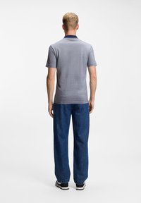 Άνδρας που στέκεται με την πλάτη στραμμένη, φοράει γκρι κοντομάνικη πόλο μπλούζα, μπλε τζιν και μαύρα και λευκά αθλητικά παπούτσια σε λευκό φόντο.