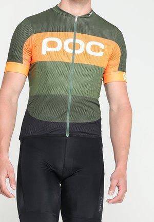Homme portant un maillot de cyclisme POC vert et orange et un cuissard rembourré noir, debout les bras détendus.