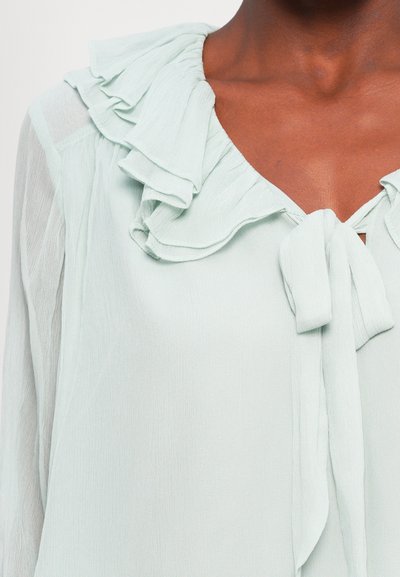 Light mint green blouse with a ruffled neckline, tie detail at the front, and sheer sleeves. Textured fabric with soft drape.