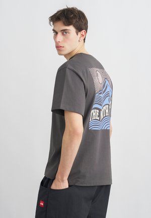 Jonge man met kort donker haar, gekleed in een grijs t-shirt met print en zwarte broek, kijkt over zijn schouder tegen een effen achtergrond.