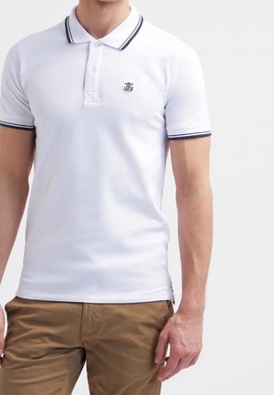 Uomo che indossa una polo bianca aderente con rifiniture nere sul collare e sulle maniche, abbinata a pantaloni marrone chiaro, in piedi su uno sfondo bianco.