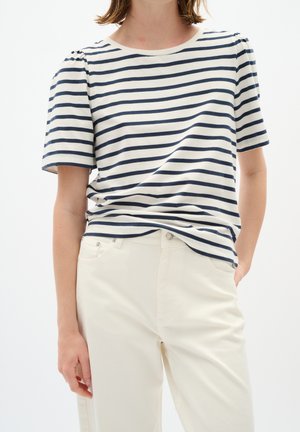 Persoon die een wit en marineblauw gestreept T-shirt met korte mouwen draagt, ingestopt in een witte broek met hoge taille, met één hand in de zak.