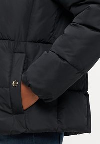 Svart puffjakke med quiltet design, overdimensjonerte ermer og en sidelomme med trykknapplukking. Glatt tekstur og varmt isolasjon.