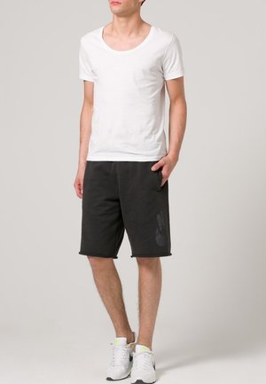 T-shirt blanc à manches courtes, col rond, associé à un short de sport noir avec poches avant et un discret logo imprimé. Chaussures de sport blanches.
