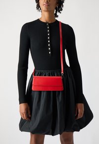 Zwarte geribbelde trui met knoopsluiting en lange mouwen, gecombineerd met een zwarte pofrok. Een rode leren crossbody tas rust op de taille.
