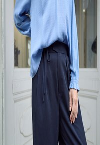 Light blue textured blouse with elastic cuffs paired with dark navy high-waisted trousers featuring a tied waist.
