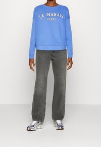Sudadera de cuello redondo azul con "LE MARAIS PARIS" en letras blancas, combinada con jeans rectos grises y zapatillas deportivas multicolores.