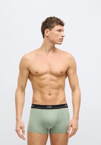 Zielone bawełniane bokserki z czarnym pasem z logo, pokazane na męskim modelu w profilu bocznym na neutralnym tle.