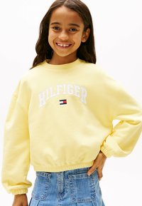 Sudadera amarilla con cuello redondo, puños elásticos y dobladillo. Presenta el texto "HILFIGER" en blanco y un pequeño logotipo de acento. Combinada con denim.
