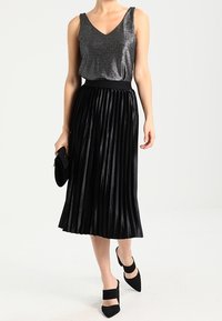 Schwarzer plissierter Rock mit tailliertem Bund, kombiniert mit einem metallischen V-Ausschnitt-Oberteil. Accessoires umfassen eine kleine schwarze Clutch und spitze Schuhe.