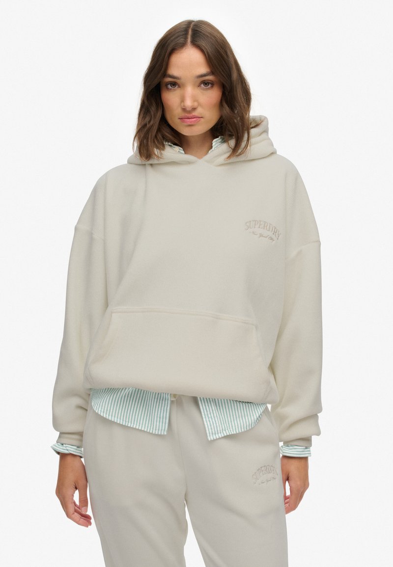 Felpa beige chiaro in tessuto morbido con una tasca frontale, caratterizzata da un piccolo logo "Superdry" ricamato sul petto. Vestibilità oversize.