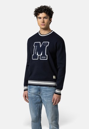 Junger Mann, der einen marineblauen Pullover mit weißen Streifen und einem großen "M"-Logo trägt, kombiniert mit hellblauen Jeans, steht vor einem einfarbigen Hintergrund.