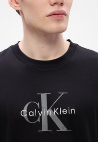 Male wearing black crew neck t-shirt with large gray "cK" and white "Calvin Klein" logo on chest, against plain light background.