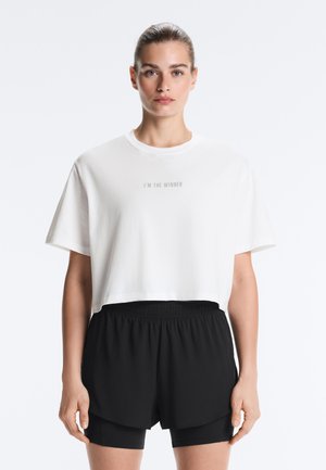 Giovane donna che indossa una maglietta bianca corta e larga con la scritta "I'M THE WINNER" e pantaloncini sportivi neri a più strati, guarda avanti.