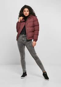 Donna con capelli ricci, che indossa un giubbotto imbottito bordeaux, un maglione a collo alto nero, jeans grigi effetto acid wash e scarpe da ginnastica alte nere, in posa con convinzione.