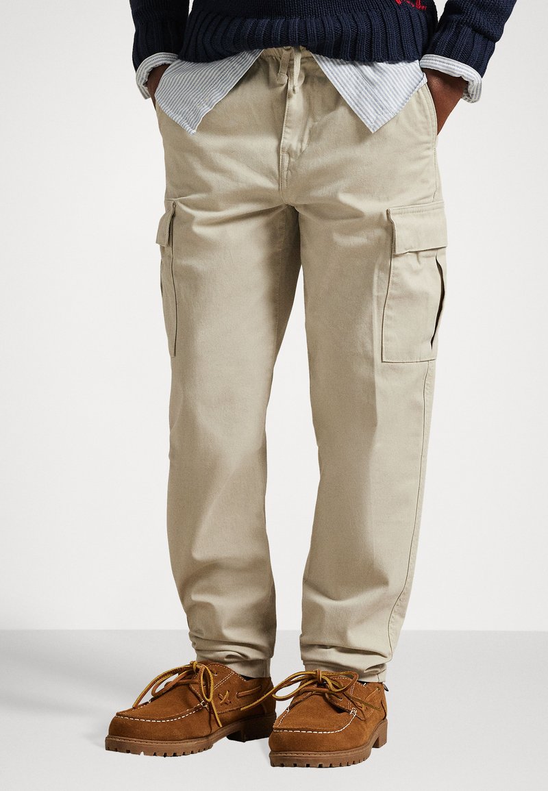 Μπέζ cargo παντελόνι με πολλαπλές πλευρικές τσέπες, κορδόνι στη μέση και άνετη εφαρμογή, συνδυασμένο με καφέ σουέτ παπούτσια με κορδόνια.