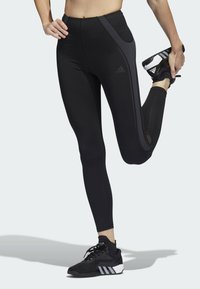Schwarze Sportleggings mit einem hohen Bund, versehen mit grauen Seitenpaneelen und reflektierenden Akzenten. Kombiniert mit schwarzen Sportschuhen.