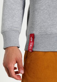 Un sweat-shirt gris avec des poignets et un ourlet côtelés, arborant une étiquette rouge avec le texte "REMOVE BE". La texture du tissu semble douce et décontractée.