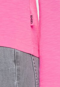 Nahaufnahme eines rosa Hemdsaums mit einem "SENSES"-Label neben der Naht einer grauen Hose, das die Textur und die Nähdetails hervorhebt.