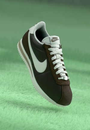 Brązowo-biały sneaker Nike z białymi sznurowadłami, widoczne logo na boku i języku, prezentowany na miękkiej zielonej powierzchni.