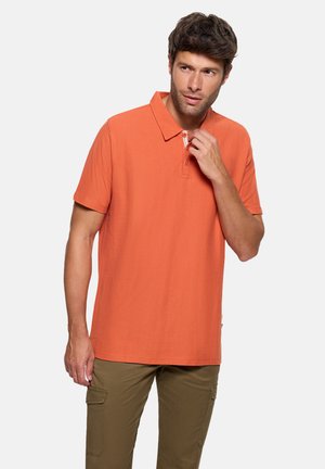 Hombre ajustándose el cuello de una camiseta polo naranja de manga corta, con pantalones cargo oliva, de pie frente a un fondo liso de color gris claro.