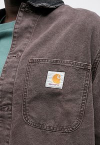 Hnědá denimová košile s černým límcem, na náprsní kapse má čtvercové logo s oranžovým motivem.