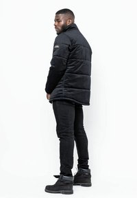 Zwarte gewatteerde jas met een hoge kraag, een gegarneerde textuur en een logo-embleem aan de zijkant. Draagt met zwarte slim-fit jeans en zwarte laarzen.