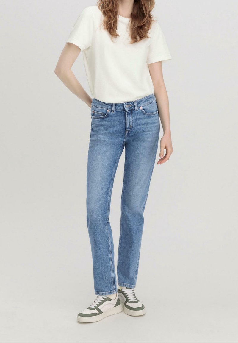 Femme portant un t-shirt blanc uni à manches courtes, un jean bleu à jambes droites et des baskets blanches avec des accents verts, debout avec une main sur la hanche.