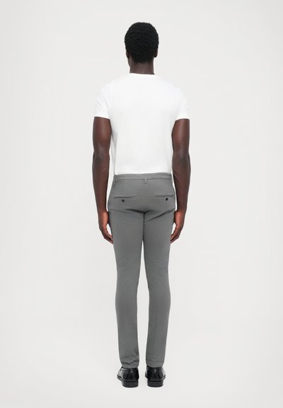 T-shirt bianco a maniche corte, pantaloni grigi slim-fit e scarpe nere, visti da dietro. I pantaloni presentano due tasche posteriori con chiusure a bottone.