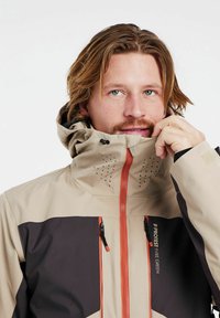 Beige en donkerbruin jack met oranje ritsen, hoge kraag en geperforeerde details op de capuchon. Bevat zwarte accenten en logo's.