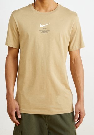 Mann trägt beige Nike-T-Shirt mit weißem Logo und Schriftzug, kombiniert mit dunkelgrünen Cargo-Shorts, steht vor einem schlichten hellen Hintergrund.