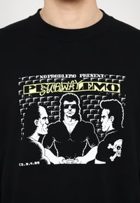 Black cotton t-shirt featuring retro graphic print of three characters in a cartoon style, with yellow and white text and skull design detail.