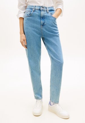 Persoon draagt lichtblauwe high-waist jeans, witte sneakers en een wit lang shirt met één hand in de zak.