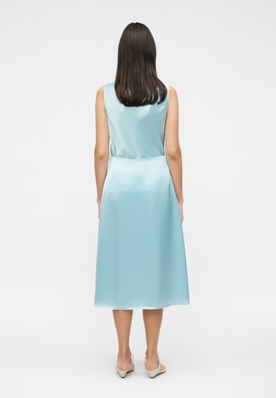 Γυναίκα με ίσια καστανά μαλλιά, φορώντας μια αμάνικη, ανοιχτό μπλε σατέν midi φόρεμα και μπεζ ψηλοτάκουνα, στέκεται με την πλάτη σε λευκό φόντο.