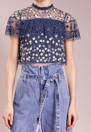 Marineblauwe doorschijnende crop top met meerkleurige bloemenborduursels, gecombineerd met lichtblauwe high-waisted jeans met riem.