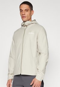 Lys beige udendørs jakke med lynlås foran, hætte og logo på brystet. Har et strømlinet design og struktureret stof.