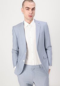 Traje azul claro a medida, confeccionado en una tela suave con solapa de muesca, combinado con una camisa blanca de botones; presenta una textura sutil y bolsillos.