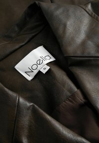 Barna bőrujjú viselet, puha tapintással, fehér címkével, amelyen az "Noella" felirat és az "S" méret szerepel, sima, elegáns kialakítással.