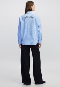 Blauw gestreepte oversized blouse met lange mouwen, met een geborduurde "IPK collectie" op de achterkant, gecombineerd met zwarte broek met wijde pijpen.