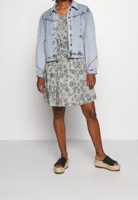 Veste en denim à manches longues sur une robe légère à motifs floraux gris clair. Sandales à plateforme noires avec des détails tissés.