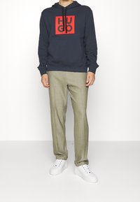 Persona vistiendo una sudadera navy con el logo rojo de HUGO, pantalones beige y zapatillas blancas, de pie contra un fondo blanco liso.