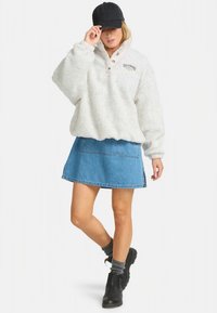 Kvinna iklädd en vit fleecetröja, jeanskjol, svart keps, grå strumpor och svarta ankelboots, poserar med en hand på kepsen.