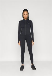 Melnas sporta komplekts, kas sastāv no ilgu piedurkņu pieguļošas blūzes ar augstu apkakli un rāvējslēdzēju, kas kopā ar atbilstošiem legingiem un melniem sporta apaviem.