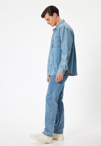 Chaqueta de denim azul claro con cierre de botones y bolsillo, combinada con jeans azul claro a juego y zapatillas blancas. Diseño simple y clásico.