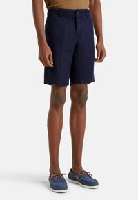 Námorně modré lněné šortky s plochým předním dílem, na knoflík a s bočními kapsami. Nosené s modrými námořnickými botami a hnědým tričkem.