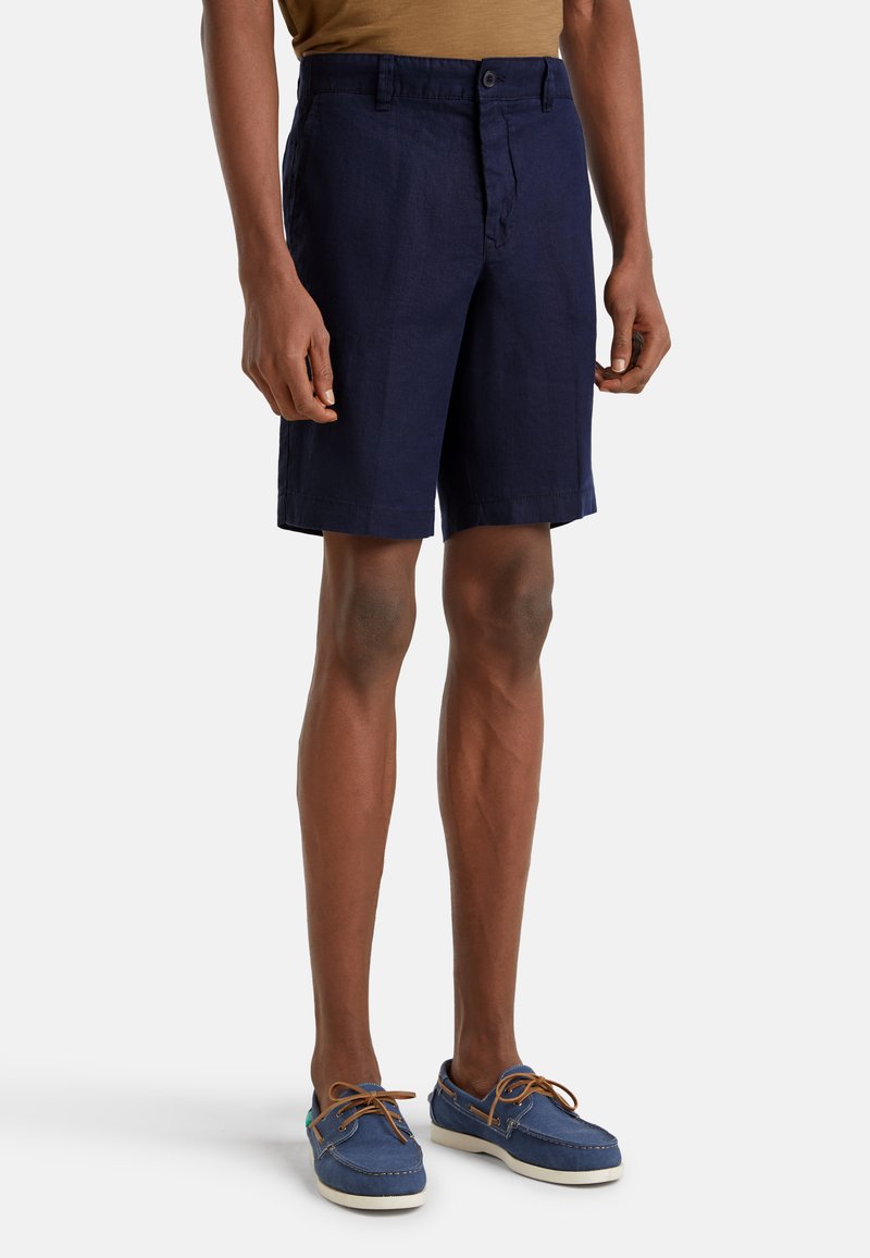 Námorně modré lněné šortky s plochým předním dílem, na knoflík a s bočními kapsami. Nosené s modrými námořnickými botami a hnědým tričkem.